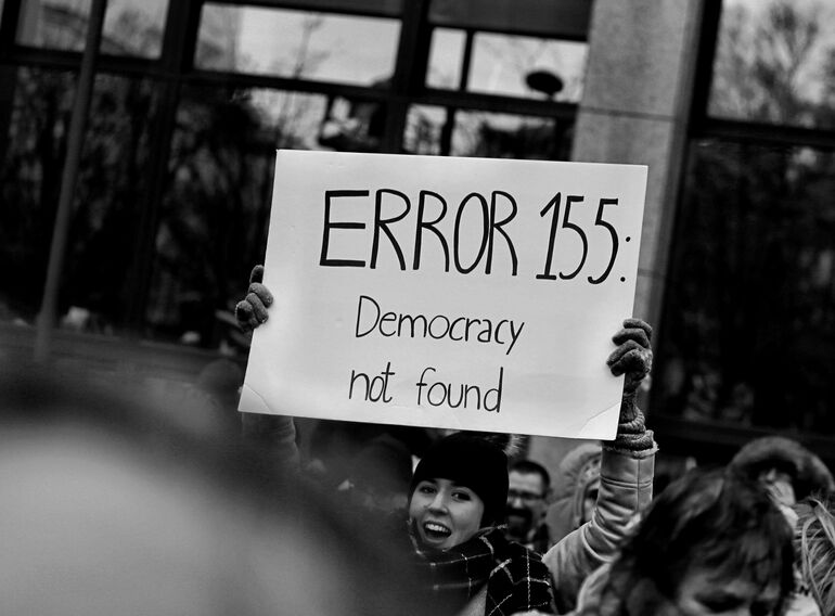 Person at a demonstration holding a sign reading 'ERROR 155: democracy not found' - Cookiebot